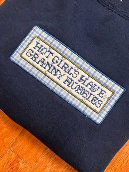 A close-up of a navy blue sweatshirt featuring a rectangular embroidered patch with a blue and white gingham border and the text 'HOT GIRLS HAVE GRANNY HOBBIES' in blue lettering on a cream background.